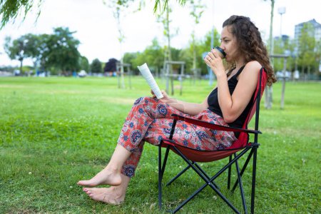 Téléchargez les photos : Young woman sitting on camping chair and reading a book - en image libre de droit