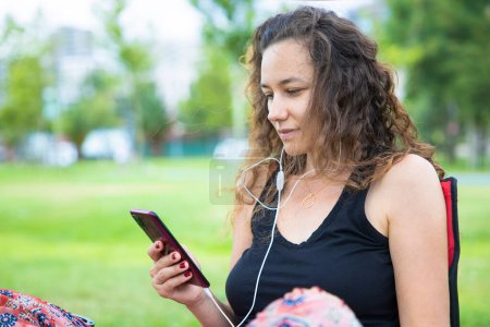 Photo for Young woman is listening to music through a pair of white earphones. She is sitting on a camping chair in a par - Royalty Free Image