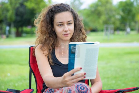 Foto de Mujer joven sentada en una silla de camping y leyendo un libro - Imagen libre de derechos
