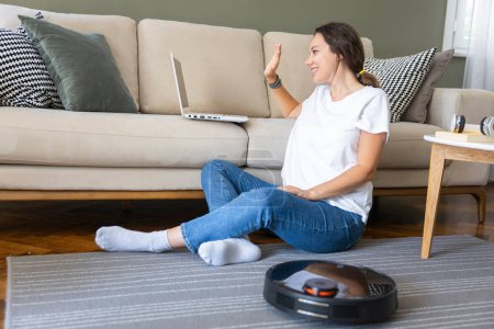 Foto de Robot aspiradora de limpieza de la sala de estar. Joven mujer disfrutar del descanso, sentado en el sofá en casa - Imagen libre de derechos