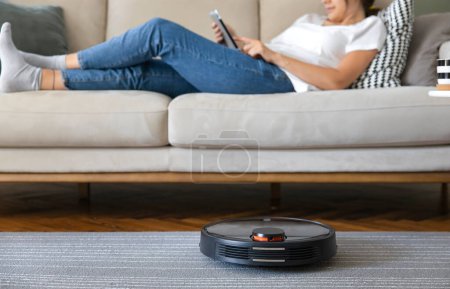 Photo for Robot vacuum cleaner cleaning the living room. Young woman enjoy rest, sitting on sofa at home - Royalty Free Image