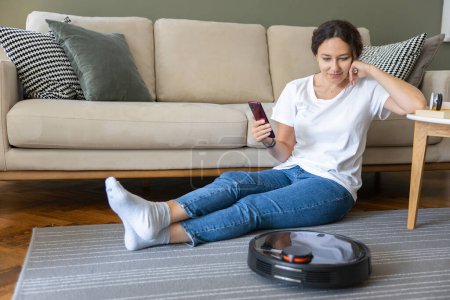 Foto de Robot vacuum cleaner cleaning the living room. Young woman enjoy rest, sitting on sofa at home - Imagen libre de derechos