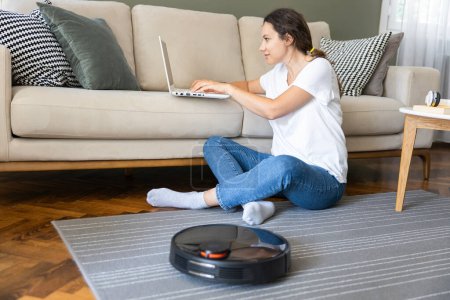 Téléchargez les photos : Robot vacuum cleaner cleaning the living room. Young woman enjoy rest, sitting on sofa at home - en image libre de droit