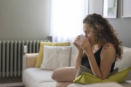 Téléchargez les photos : Jeune femme assise dans un canapé près de la fenêtre avec une tasse de café chaud - en image libre de droit