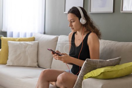 Téléchargez les photos : Jeune femme assise sur le canapé dans le salon, utilisant un téléphone intelligent et écoutant de la musique - en image libre de droit