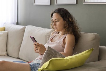 Téléchargez les photos : Jeune femme vérifiant les médias sociaux tenant smartphone à la maison. Utilisation de téléphone mobile app shopping en ligne, commander la livraison se détendre sur le canapé - en image libre de droit