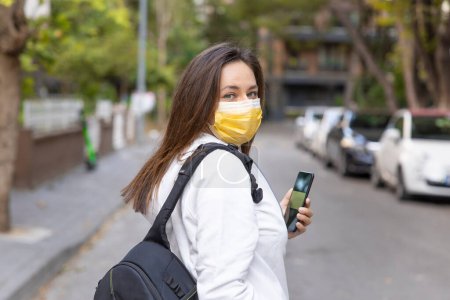 Photo for Close-up of young woman in protective mask using smartphone. Brunette woman standing on city street reading news about coronaviru - Royalty Free Image