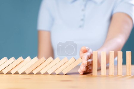 Woman hand stopping falling wooden dominoes effect on wooden table