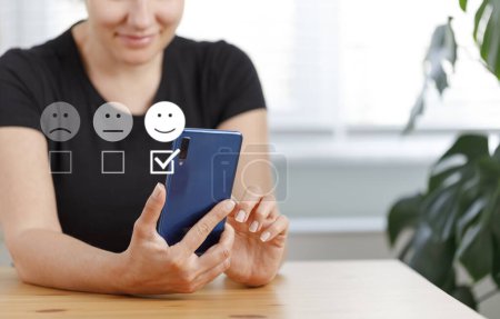 Foto de Mujer joven sonriente usando un teléfono inteligente y ella está presionando emoticono cara sonriendo en satisfacción en el tacto virtual. Concepto de evaluación del servicio al cliente. - Imagen libre de derechos