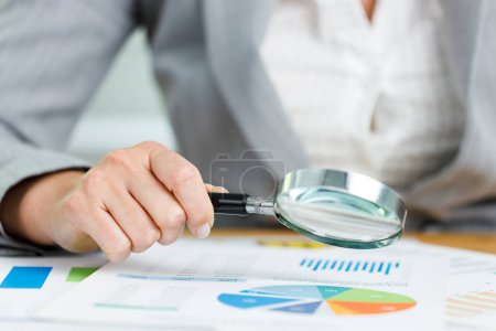 Photo for Business woman holding a magnifying glass with graph information document on the desk - Royalty Free Image