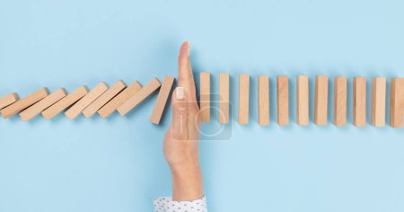 Woman hand stopping falling wooden dominoes effect on blue solid ground
