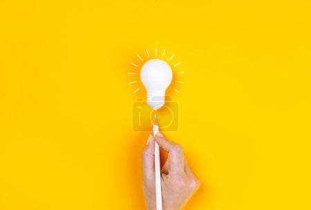 Téléchargez les photos : Main féminine pointant vers une ampoule avec stylo blanc. Créativité d'entreprise et concepts d'inspiration sur fond jaune. - en image libre de droit
