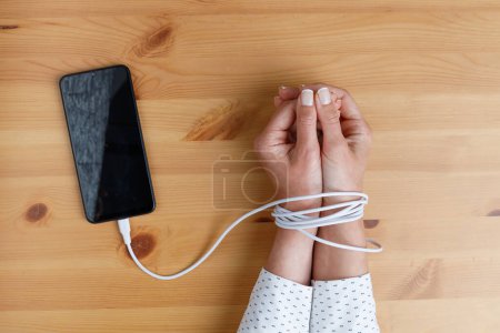 Photo for Woman's hands wrapped on wrists with mobile phone cable as handcuffs. Addiction to social networks - Royalty Free Image