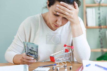 Photo for Frustrated young woman planning monthly expenses holding scanty amount of money. Empty shopping basket in front. High prices and rising inflation. Problem of families and household budget - Royalty Free Image