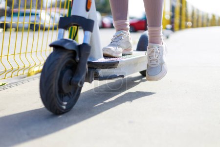 Foto de Acercamiento en piernas de mujer pies de pie en el scooter de empuje eléctrico en el pavimento con zapatillas de deporte en el día de verano - Imagen libre de derechos