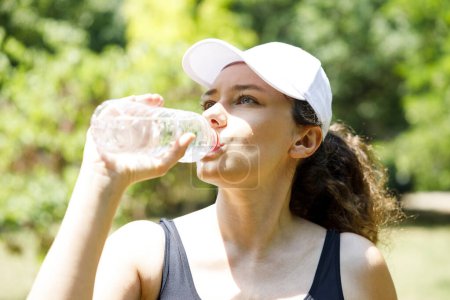 Foto de La joven deportista está bebiendo agua entre trotar. Agua potable femenina caucásica después de ejercicios o deporte. - Imagen libre de derechos