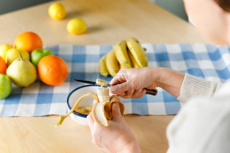 Photo for Woman sitting by the table at home cutting banana. She is preparing fruit salad at home - Royalty Free Image