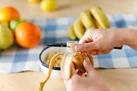 Téléchargez les photos : Femme assise à la table à la maison coupant de la banane. Elle prépare une salade de fruits à la maison - en image libre de droit