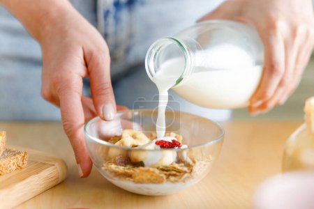 Photo for Young woman preparing healthy breakfast, pouring milk over cereals on the kitchen counter - Royalty Free Image
