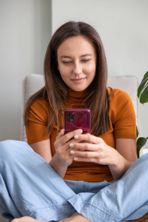 Téléchargez les photos : Jeune femme utilisant un téléphone portable tout en étant assis un canapé à la maison - en image libre de droit