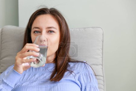 Téléchargez les photos : Une jeune femme buvant de l'eau assise sur une chaise à la maison. Avantages pour la santé de boire suffisamment d'eau concepts. - en image libre de droit