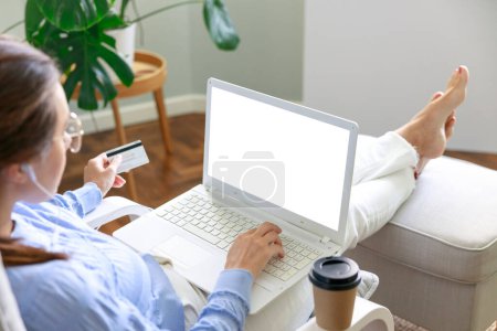 Téléchargez les photos : Jeune femme utilisant un ordinateur portable tout en étant assis à la maison et tenant une carte de crédit shopping en ligne - en image libre de droit