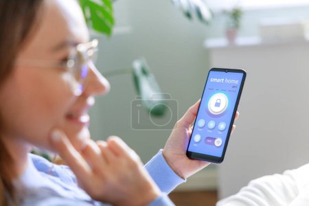 Téléchargez les photos : Jeune femme tenant le téléphone avec app maison intelligente sur l'écran dans la chambre. Concept de contrôle de la sécurité à domicile à partir d'un écart mobile - en image libre de droit