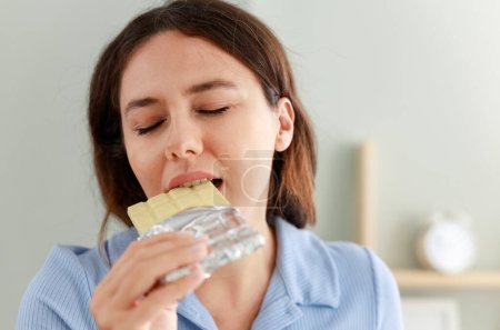 Foto de Joven alegre comiendo barra de chocolate blanco en casa - Imagen libre de derechos