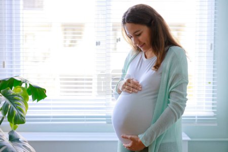 Photo for Happy pregnant young woman touching her pregnant belly and standing by the window - Royalty Free Image