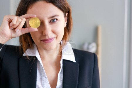 Téléchargez les photos : Souriant jeune femme couvrant l'oeil avec Bitcoin monnaie de pièce dans le bureau - en image libre de droit