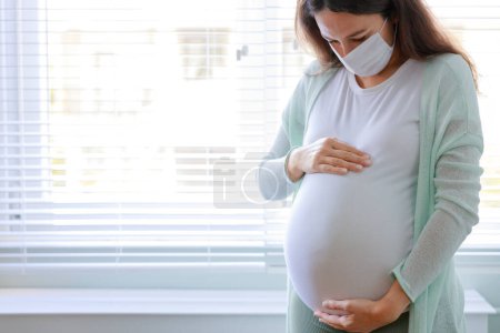Téléchargez les photos : Jeune femme enceinte en masque médical en quarantaine à la maison. Période pandémique de coronavirus à domicile - en image libre de droit