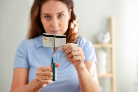 Foto de Mujer joven con expresión facial seria cortando una tarjeta de crédito con tijeras en su habitación en casa - Imagen libre de derechos