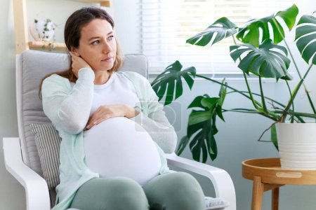 Photo for Young woman suffering from nausea and headache during pregnancy. Sitting in a comfortable armchair at home, looking unhappy and worried - Royalty Free Image