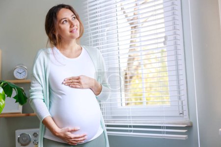 Téléchargez les photos : Joyeux enceinte jeune femme touchant son ventre enceinte et debout près de la fenêtre - en image libre de droit