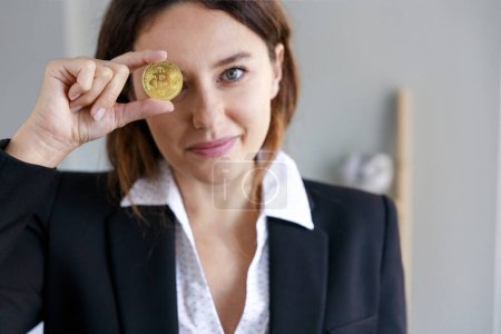 Foto de Mujer joven sonriente cubriendo el ojo con moneda bitcoin en la oficina. El foco está en la moneda - Imagen libre de derechos