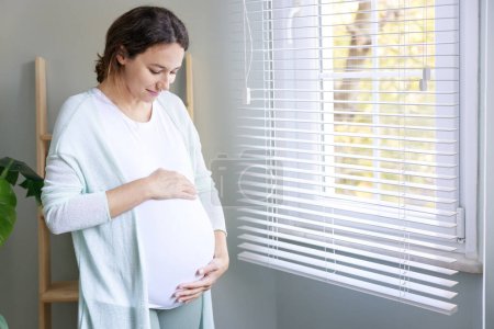 Foto de Feliz embarazada joven tocando su vientre embarazada y de pie junto a la ventana - Imagen libre de derechos