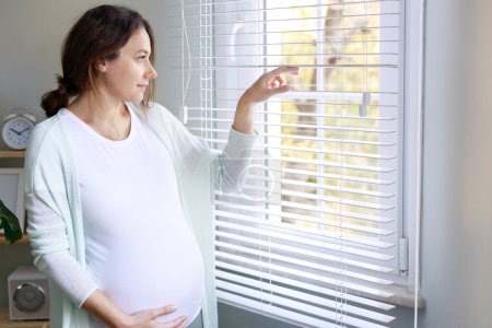 Téléchargez les photos : Joyeux enceinte jeune femme touchant son ventre enceinte et debout près de la fenêtre - en image libre de droit