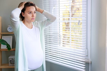 Téléchargez les photos : Jeune femme souffrant de maux de tête pendant la grossesse. Elle se tient près de la fenêtre dans sa chambre à la maison, l'air malheureuse et inquiète - en image libre de droit