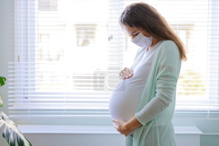 Foto de Mujer embarazada joven con máscara médica en cuarentena en casa. Período pandémico del coronavirus en casa - Imagen libre de derechos