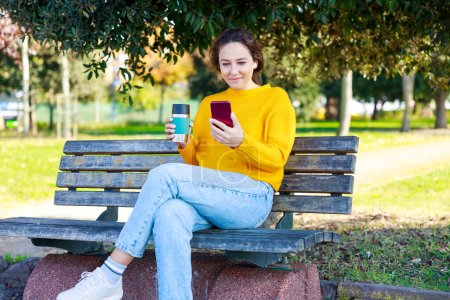 Foto de Mujer joven se relaja en el descanso en el parque. Ella está tomando café en el parque y mirando su teléfono - Imagen libre de derechos
