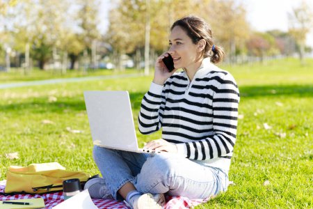 Téléchargez les photos : Jeune femme assise dans le parc avec ordinateur portable et parlant au téléphone - en image libre de droit