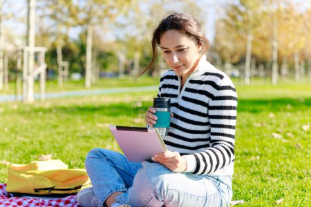 Téléchargez les photos : Jeune femme utilisant une tablette assise dans le parc. Une femme heureuse tient une tasse de café. Elle est assise sur une couverture de pique-nique - en image libre de droit