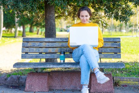 Foto de Mujer joven usando computadora portátil. La hembra feliz sostiene la taza de café. Ella está sentada en el banco - Imagen libre de derechos