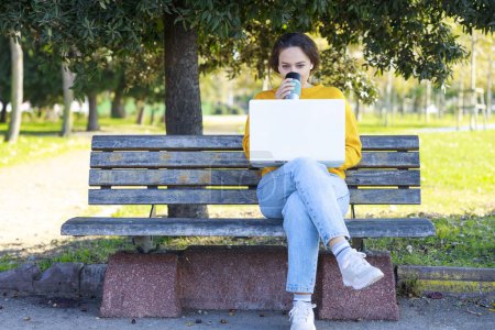 Téléchargez les photos : Jeune femme utilisant un ordinateur portable. Une femme heureuse tient une tasse de café. Elle est assise sur le banc - en image libre de droit
