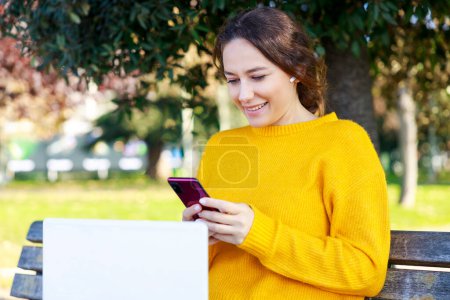 Foto de Mujer joven usando el ordenador portátil y sosteniendo el teléfono inteligente. La mujer feliz está haciendo una videollamada en el parque. Ella está sentada en el banco - Imagen libre de derechos