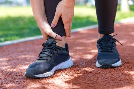 Foto de Lesión por entrenamiento: Mujer joven usa las manos en el tobillo mientras corre en el campo de atletismo - Imagen libre de derechos