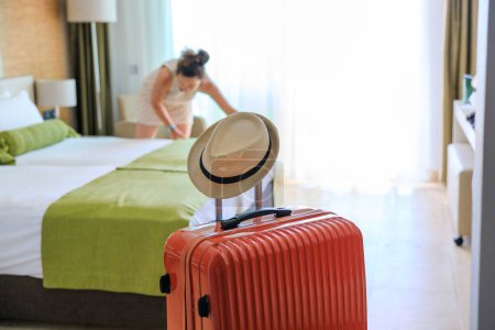 Photo for Orange color suitcase delivered in hotel room. Young woman changing bed sheets out of focus - Royalty Free Image