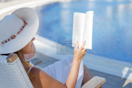 Foto de Joven mujer leyendo libro en cubierta silla cerca de la piscina - Imagen libre de derechos