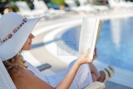Photo for Young woman reading book in deck chair near swimming pool - Royalty Free Image