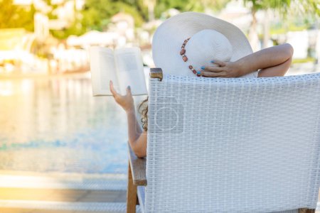 Young woman reading book in deck chair near swimming pool